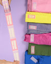 A person's hands hold a long strip of pink and lavender fabric labels next to a stack of folded colorful fabrics in pink, yellow, gray, green, and blue, all featuring matching labels on a solid lavender background.