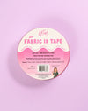 A round spool of KATM Mini Fabric ID Tape on a solid pastel pink background. The tape is packaged in a pink and white circular label with product details, a barcode, and a small illustration of a woman sewing.