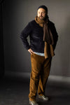 Man wearing a dark sweater, brown scarf, and brown The Elling Men’s Trouser against a grey background