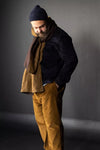 Man wearing a dark jacket, brown scarf, and mustard The Elling Men’s Trouser against a gray background