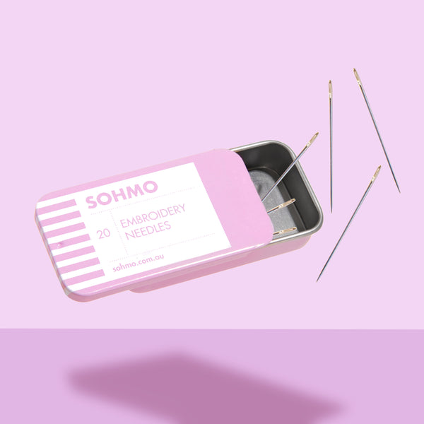 Pink tin box labeled 'SOHMO' with embroidery needles on a pink background