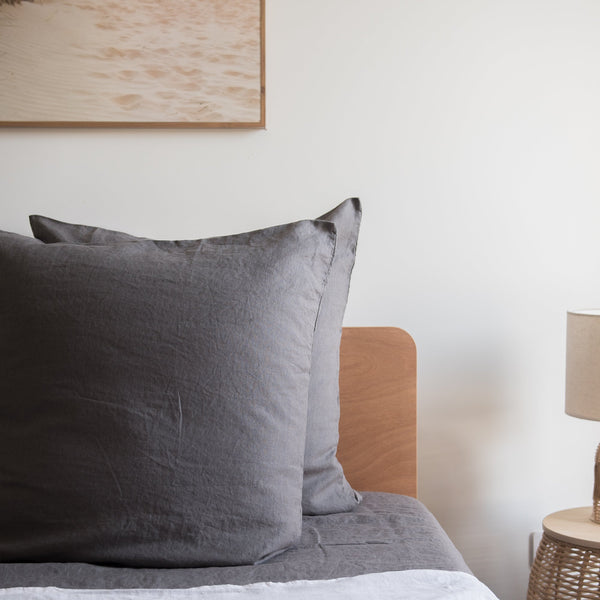 Charcoal grey European linen pillows on a bed with a wooden headboard and side table in the background.