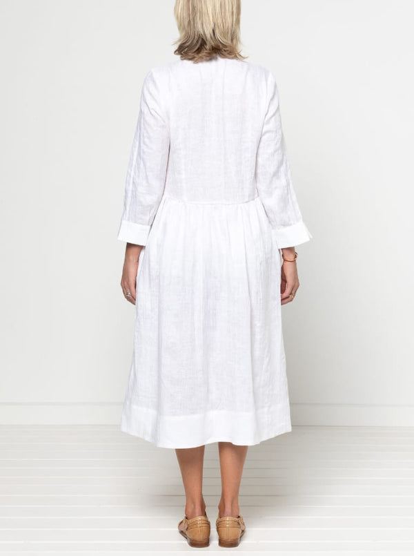 Person wearing a white dress against a plain background -Hampton Woven Dress Sewing Pattern – Mid-calf shirtdress with gathered skirt, curved waist, sleeve or sleeveless options, and inseam pockets.