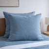 Two Lake Blue 100% Linen European Pillowcase on a bed with a lamp and wooden headboard in the background.