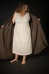 Woman holding a brown coat over a white dress made using The Lilian Sewing Pattern by Merchant & Mills against a black background