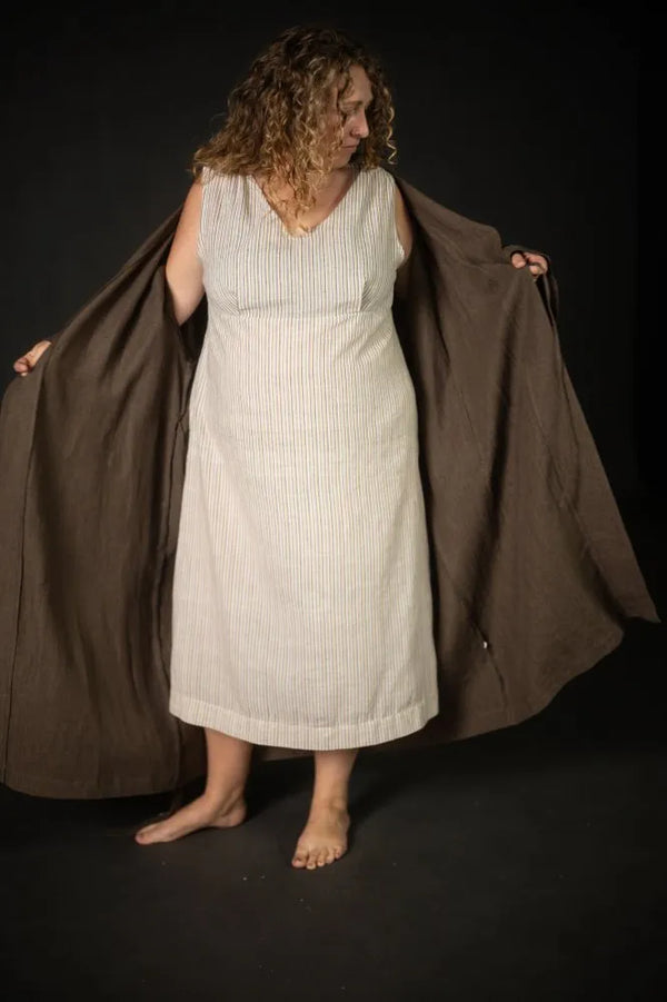 Woman holding a brown coat over a white dress made using The Lilian Sewing Pattern by Merchant & Mills against a black background