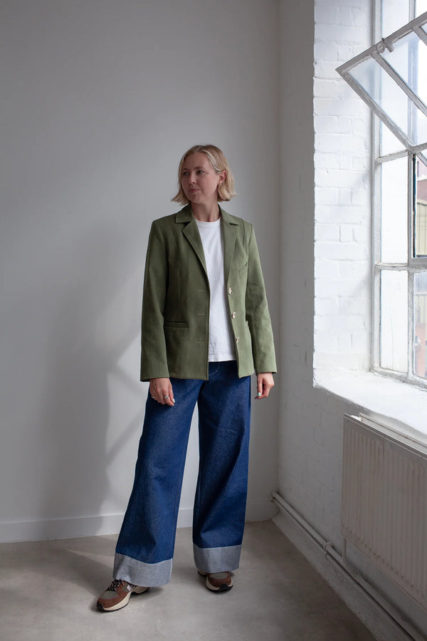 Person wearing a green jacket made using Naomi Blazer PDF Sewing Patternand blue jeans standing in a room with a large window.