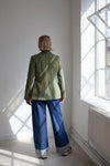 Person wearing a green jacket made using Naomi Blazer PDF Sewing Patternand blue pants standing in a room with sunlight streaming through a window.