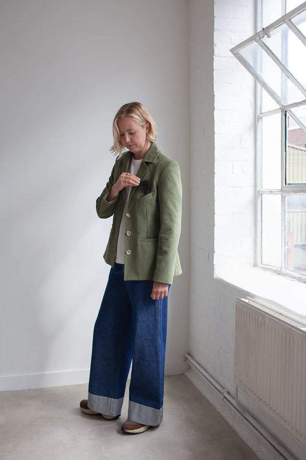 Person wearing a green jacket made using Naomi Blazer PDF Sewing Patternand blue pants standing in a room with a window.