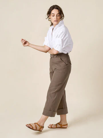 Woman wearing a white shirt and brown pants made of Rika Pants sewing pattern in cropped tailored style with hem cuffs and tabbed waistbandon a beige background