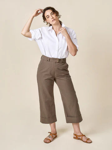 Woman wearing a white shirt and brown pants made of Rika Pants sewing pattern in cropped tailored style with hem cuffs and tabbed waistband against a beige background