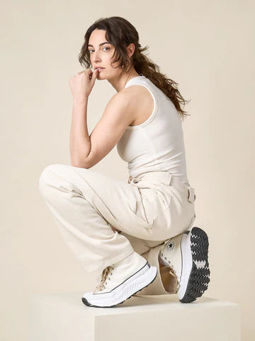 Woman in beigejumpsuit and black sneakers sitting on a white surface with a beige background