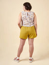 Woman wearing a sleeveless floral top and mustard yellow shorts made using Sepia Pants and Shorts sewing patternon a beige background