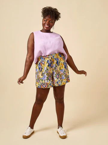 Woman wearing a pink sleeveless top and colourful floral shorts made using Sepia Pants and Shorts sewing patternon a beige background