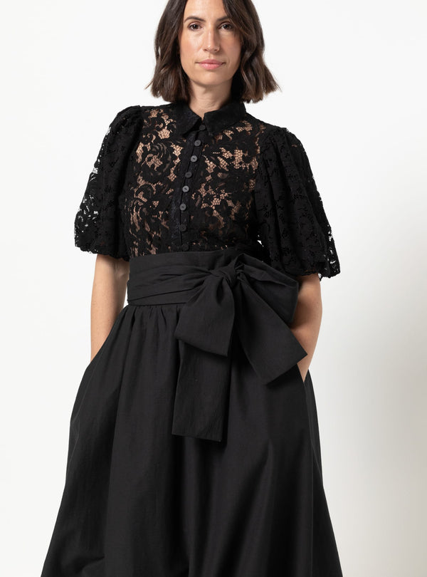 A woman wearing a black lace blouse with short puff sleeves and a high collar, paired with a black skirt made using Sophia Woven Bubble Skirt and Pant sewing pattern featuring a large waist bow, photographed against a plain light background.