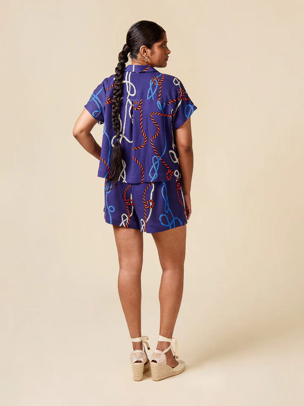 A woman is shown from the back wearing a matching purple two-piece set with a colorful chain print, consisting of a short-sleeve top and shorts, paired with beige espadrille wedge sandals against a solid beige background.