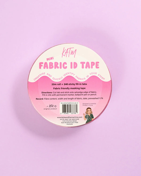 A round spool of KATM Mini Fabric ID Tape on a solid pastel pink background. The tape is packaged in a pink and white circular label with product details, a barcode, and a small illustration of a woman sewing.