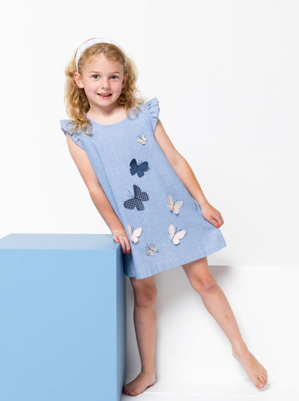 Young girl wearing a blue dress made using andie kids dress sewing patternwith butterfly patterns on a white background