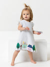 A toddler girl stands barefoot on a white studio set wearing a white ruffled dressmade using andie kids dress sewing pattern with a colorful house and tree print near the hem, holding a toy wand.