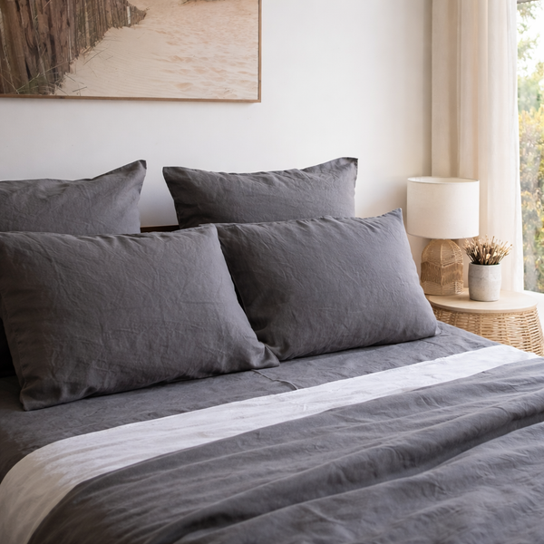 Charcoal grey linen pillowcases set on a bed with a lamp and window in the background