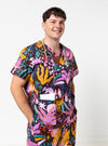 A man wearing a colorful tropical print medical scrub set, featuring a short-sleeve top and matching pants with a stethoscope around his neck, photographed against a plain white studio background.