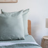 Harbour Sage European linen pillowcaseson a bed with a wooden headboard and lamp in the background.