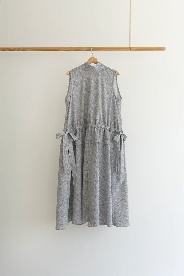 A sleeveless gray and white textured dress with side tie details, hanging on a wooden hanger from a wooden clothing rack against a plain white wall.