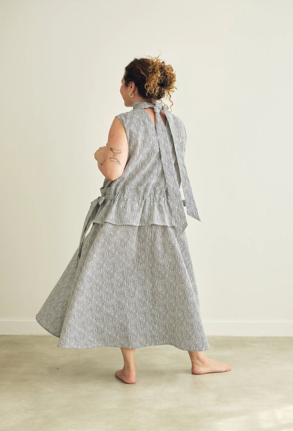 A woman wearing a sleeveless gray patterned dress with a tie-back neckline, shown from behind in a minimalist indoor setting with a light beige wall and concrete floor.