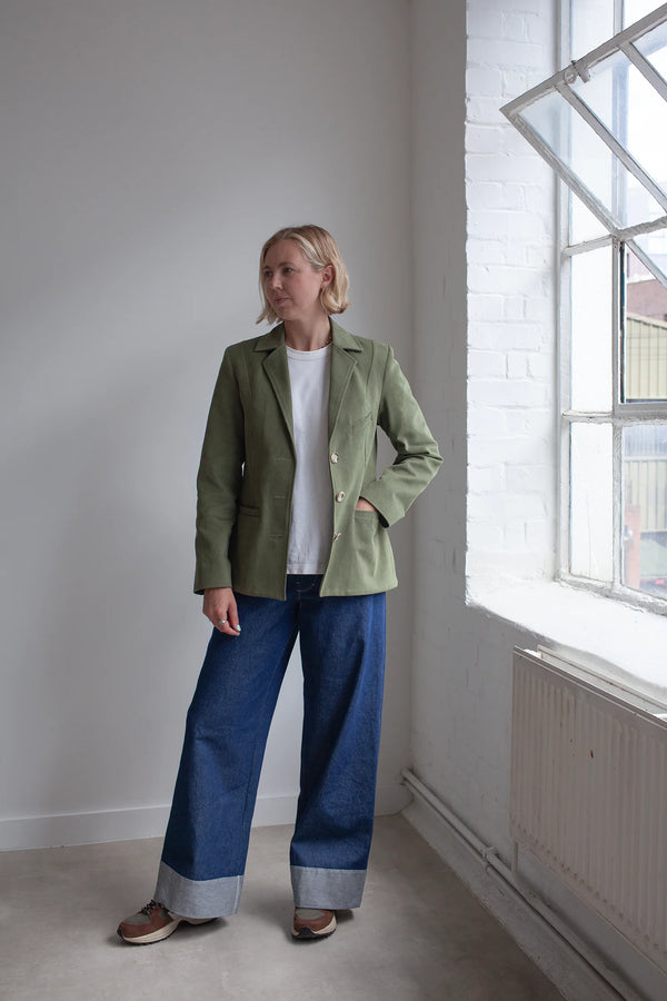 Woman wearing a green jacket made using Naomi Blazer PDF Sewing Patternand blue jeans standing in a room with a window.