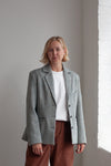 Woman wearing a gray blazer made using Naomi Blazer PDF Sewing Patternover a white shirt and brown pants against a light gray wall.