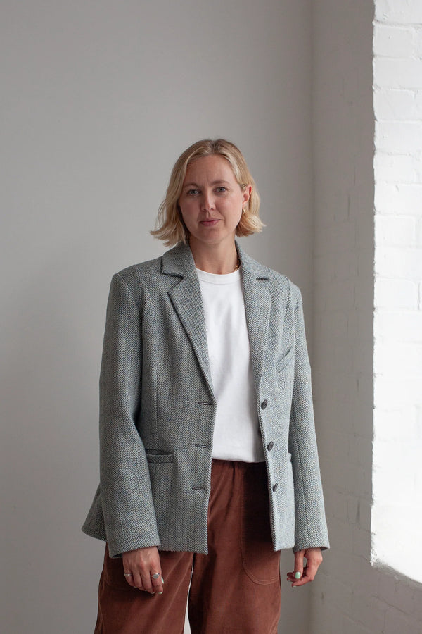 Woman wearing a gray blazer made using Naomi Blazer PDF Sewing Patternover a white shirt and brown pants against a light gray wall.