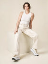 Woman wearing a white sleeveless top and beige pants sitting on a white surface with a plain background